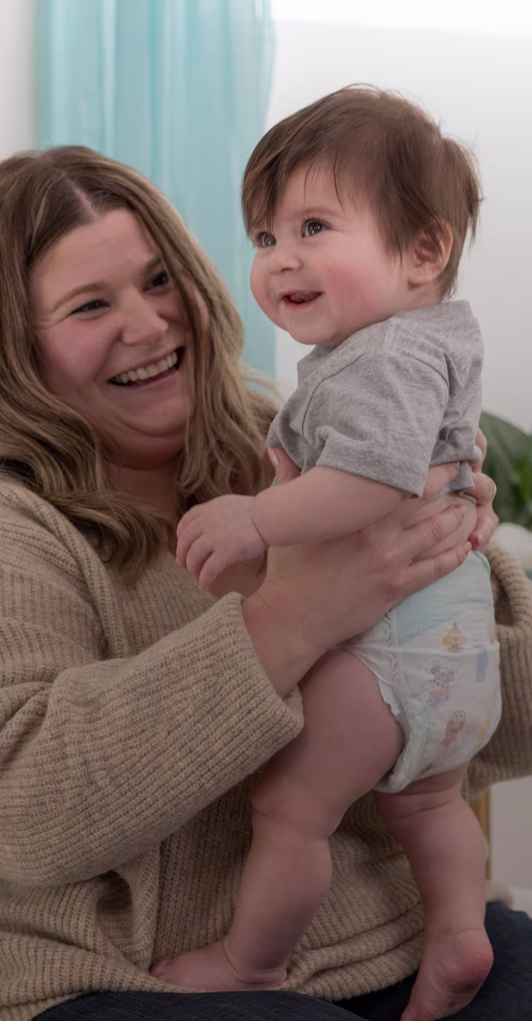 Smiling woman holds a toddler in a diaper, lifting them up as they both laugh indoors.