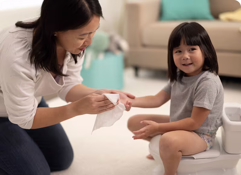 Una madre cariñosa limpia suavemente la mano de su hija con una toallita húmeda mientras ella está sentada en una bacinica, creando un momento de ternura cuando están juntas en su acogedora sala de estar.
