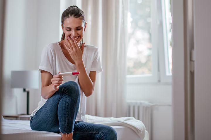 Femeie întinsă pe pat, ținând un test de sarcină într-o mână, iar cealaltă mână  o are în dreptul feței zâmbitoare.
