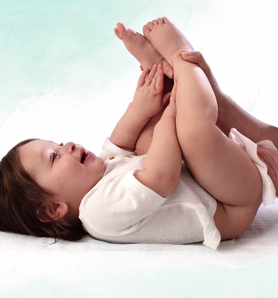 A baby lies on their back on a white surface, holding their feet up while a parent is using a wipe to clean the baby’s bottom.