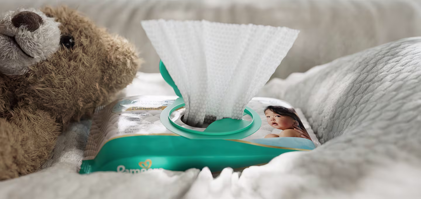 Opakowanie chusteczek dla niemowląt Pampers z jedną chusteczką wyciągniętą przez otwór, leżące na miękkim kocyku obok pluszowego misia.