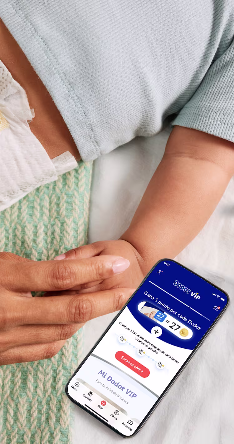 La imagen muestra un primer plano de un bebé que lleva un pañal y una camiseta azul claro, con una mano adulta que sostiene suavemente la mano del bebé. A su lado, un smartphone muestra la aplicación "Dodot VIP", que presenta un programa de fidelización para "Ganar 1 punto por cada pañal" y puntos de bonificación por escanear cada tercer pañal.