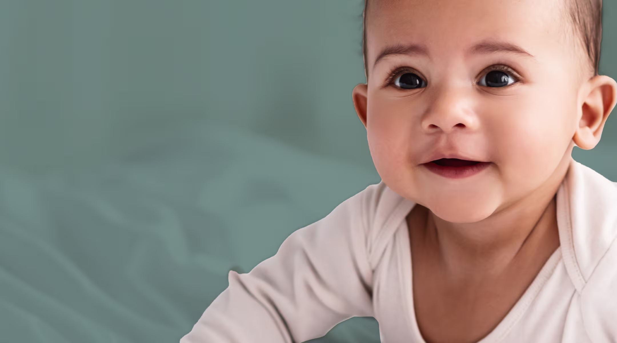 Close up of baby laying on stomach smiling and wearing a cream onesie with sage background