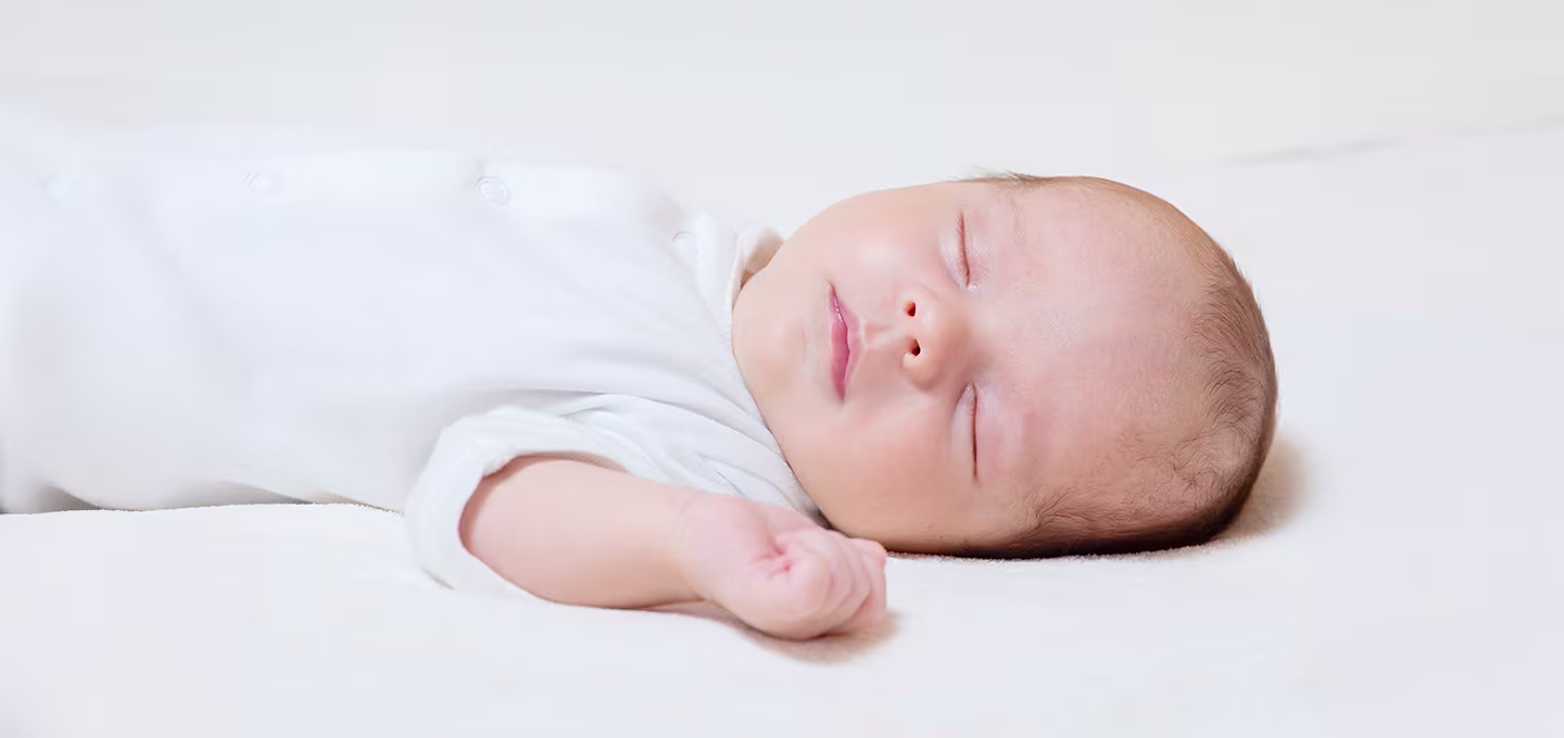 Photo d’un nouveau-né endormi sur le dos, vêtu d’un body blanc, reposant paisiblement sur un drap clair.