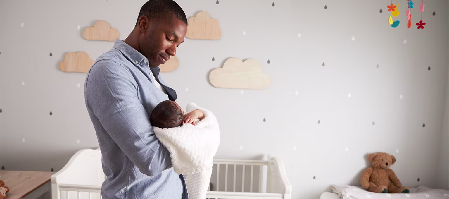 A dad carrying his newborn in a nursery room, with a crib mattress visible in the background.