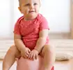 Baby in a pink polka dot outfit sitting on a potty, smiling in a bright playroom.