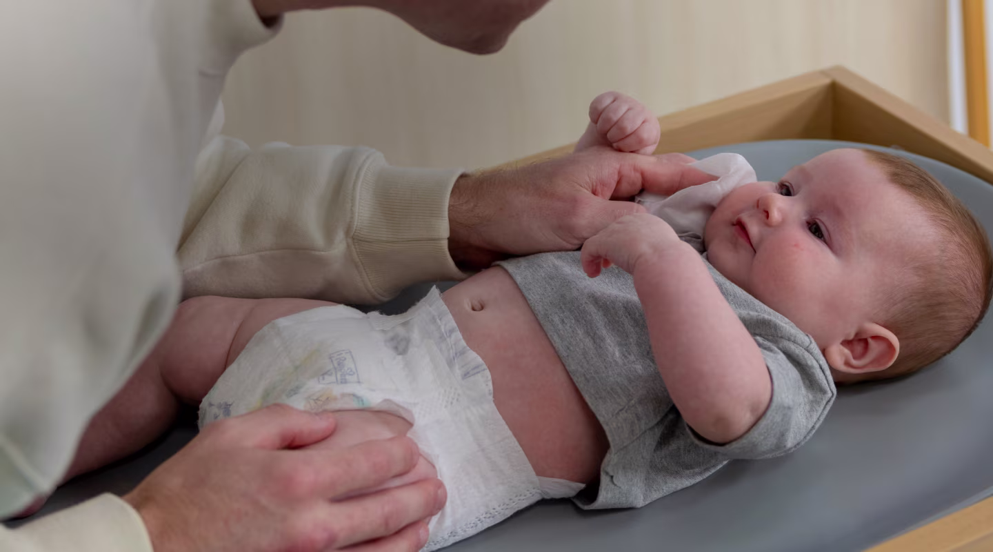 Bebé sonriente mientras está acostado en una mesa para cambiar pañales, mientras mamá le limpia suavemente la cara con una toallita húmeda Pampers.