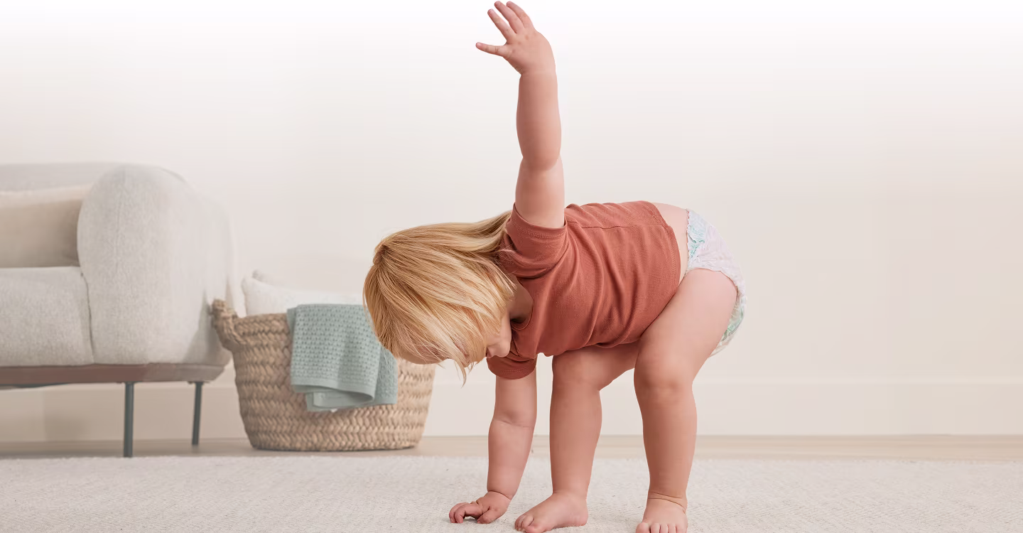 Ett litet barn går på en ljusfärgad matta framför en grå soffa.