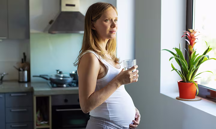 Schwangere Frau steht in der Küche und blickt aus dem Fenster, mit einem Glas Wasser in der Hand