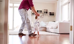 Adulto ayudando a un bebé a dar sus primeros pasos en casa, ilustrando cuándo los bebés empiezan a caminar y su desarrollo temprano.