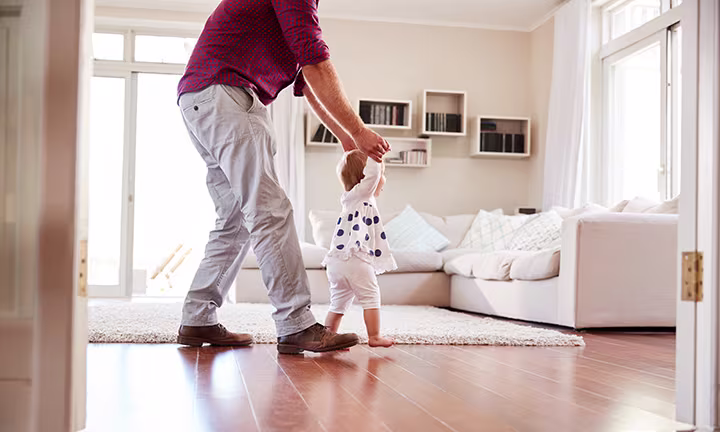 Adulto ayudando a un bebé a dar sus primeros pasos en casa, ilustrando cuándo los bebés empiezan a caminar y su desarrollo temprano.