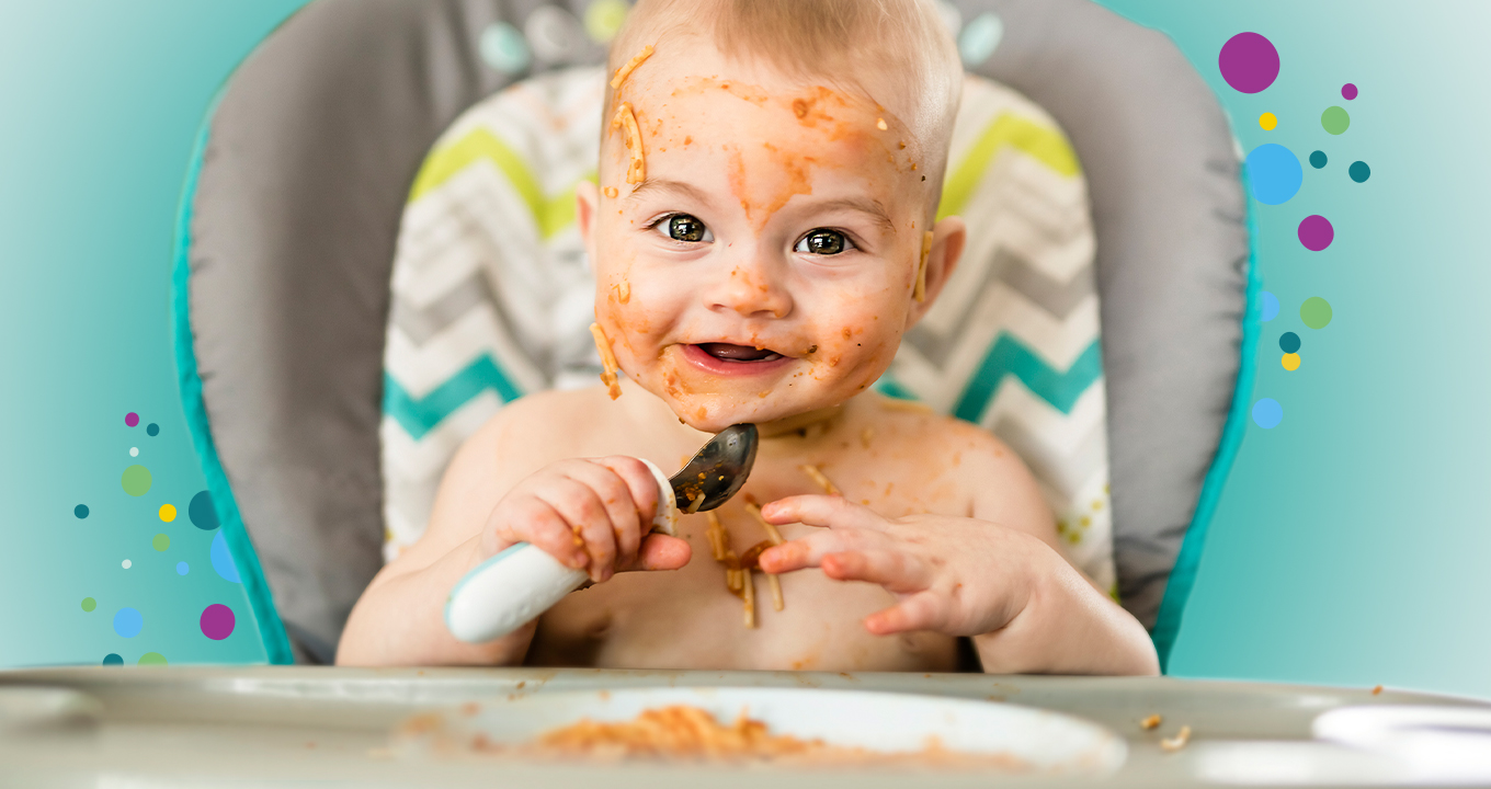 Lachendes Baby sitzt am Tisch mit Löffel in der Hand und Spaghetti mit Tomatensoße im Gesicht