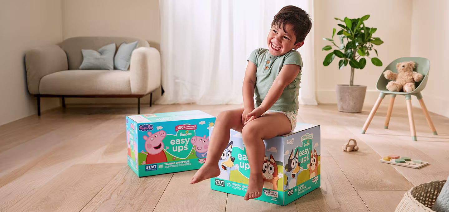 Smiling toddler sitting on Easy Ups diaper boxes in a bright playroom with plant, chair, and toys nearby