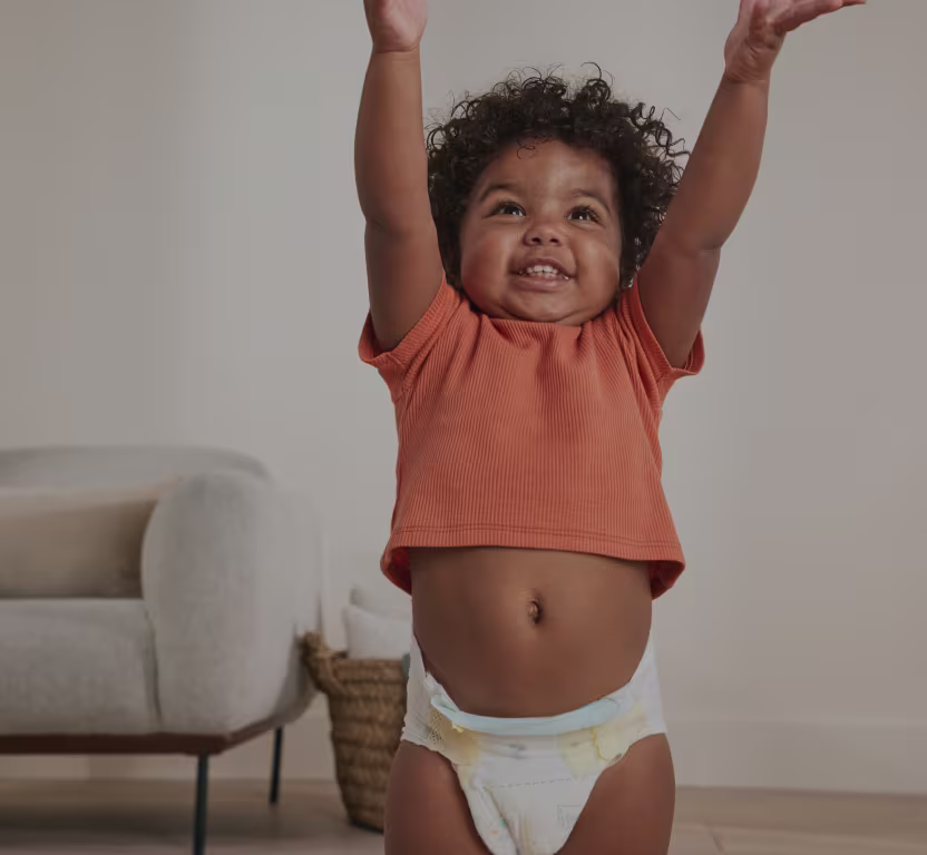 A toddler is seen in the living room smiling and stretching their arms in the air whilst wearing a tshirt and pampers diapers