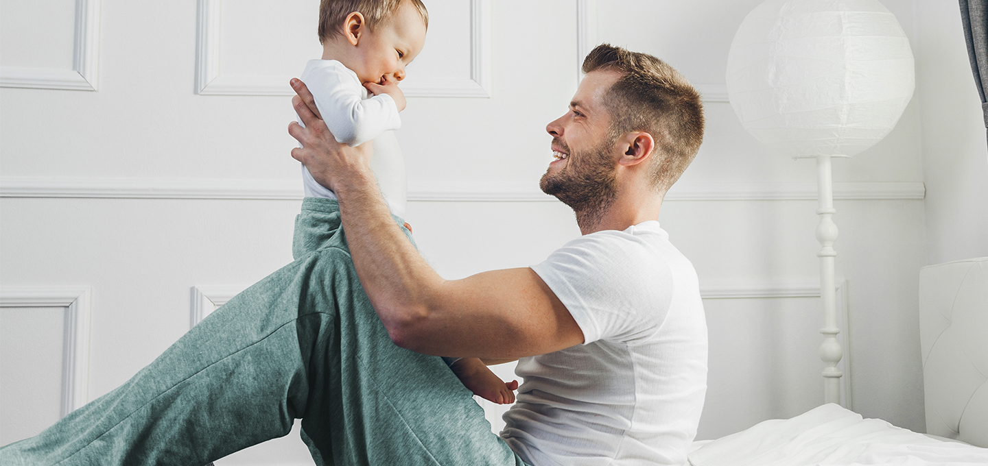 Smiling adult sits on the floor lifting a happy baby who is facing them, showing playful interaction and bonding during simple baby games.