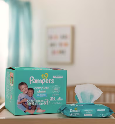 A box and individual pack of Pampers Complete Clean wipes sit on a changing table of a nursery. Showing both the Baby Fresh and unscented offerings