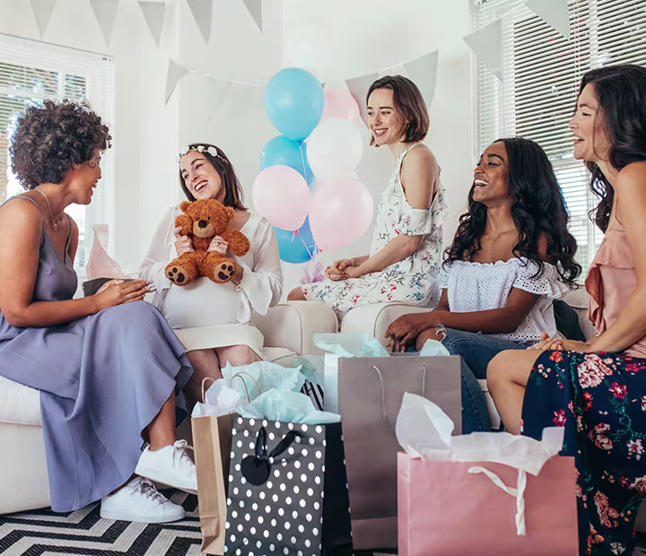 Group of women enjoying a baby shower; one holds a teddy bear surrounded by gifts and balloons.