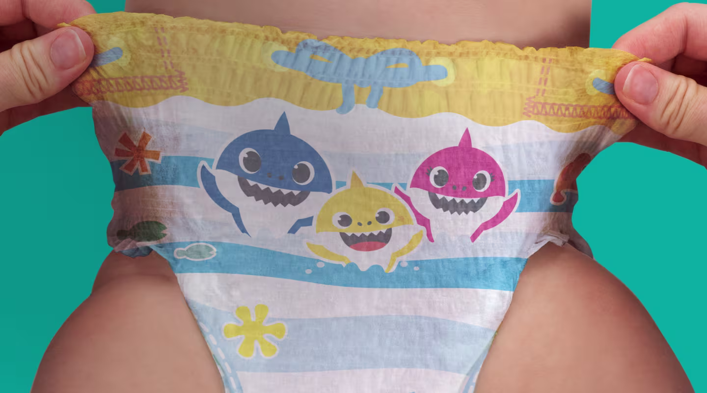 Um bebé usa uma fralda de natação com temática do Baby Shark, com riscas azuis e brancas e personagens coloridos de tubarões, enquanto as suas mãos seguram suavemente os lados.