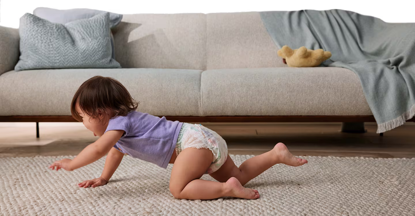 A baby crawls on a light-colored rug with a gray sofa visible in the background.
