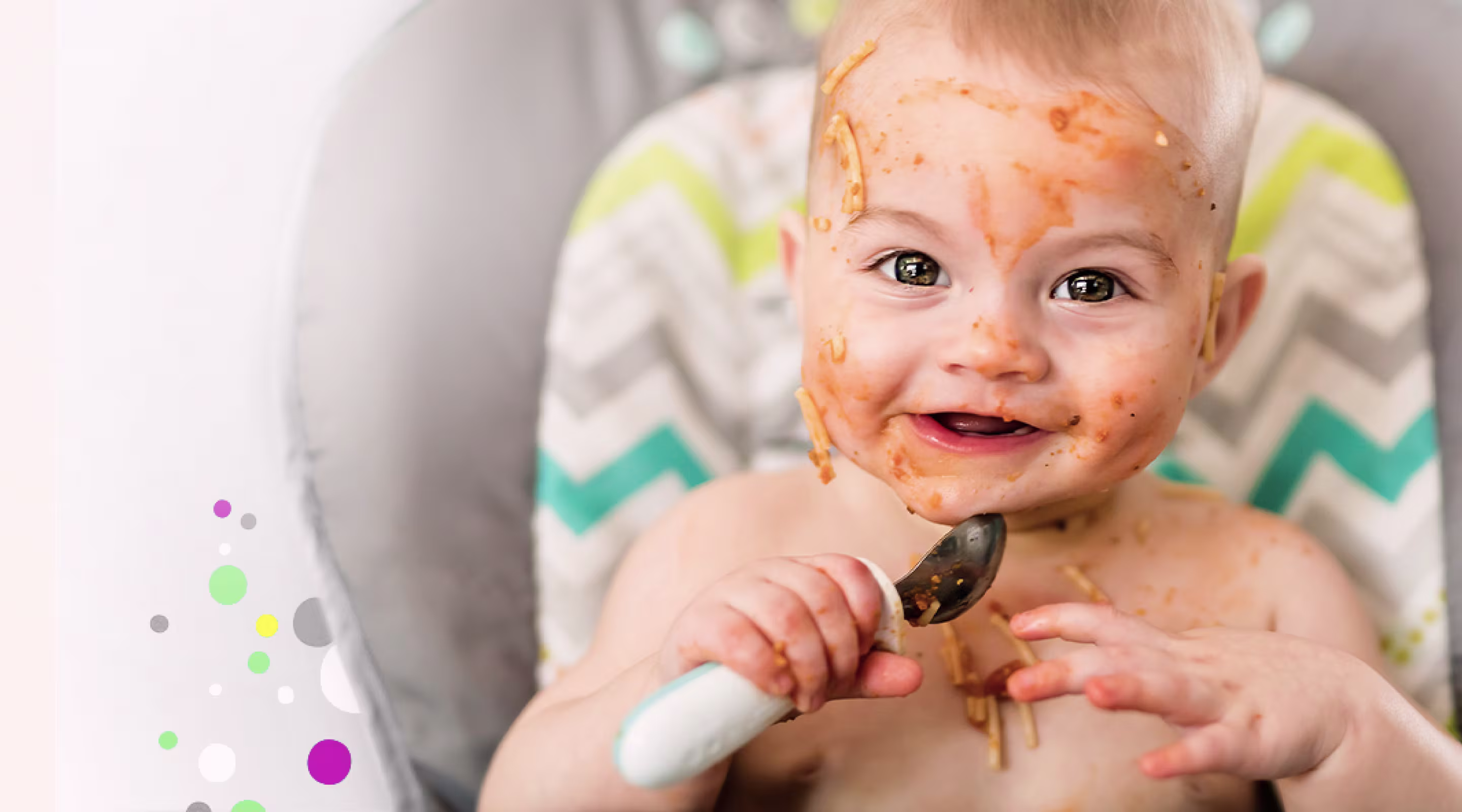 Bebé sonriente en una trona, con la cara cubierta de salsa de espagueti, sosteniendo una cuchara