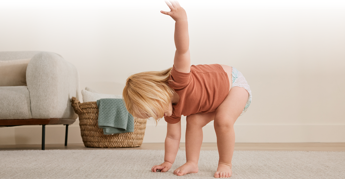A toddler walks on a light-colored rug in front of a gray sofa.