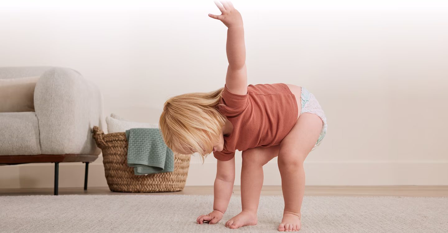 A toddler walks on a light-colored rug in front of a gray sofa.
