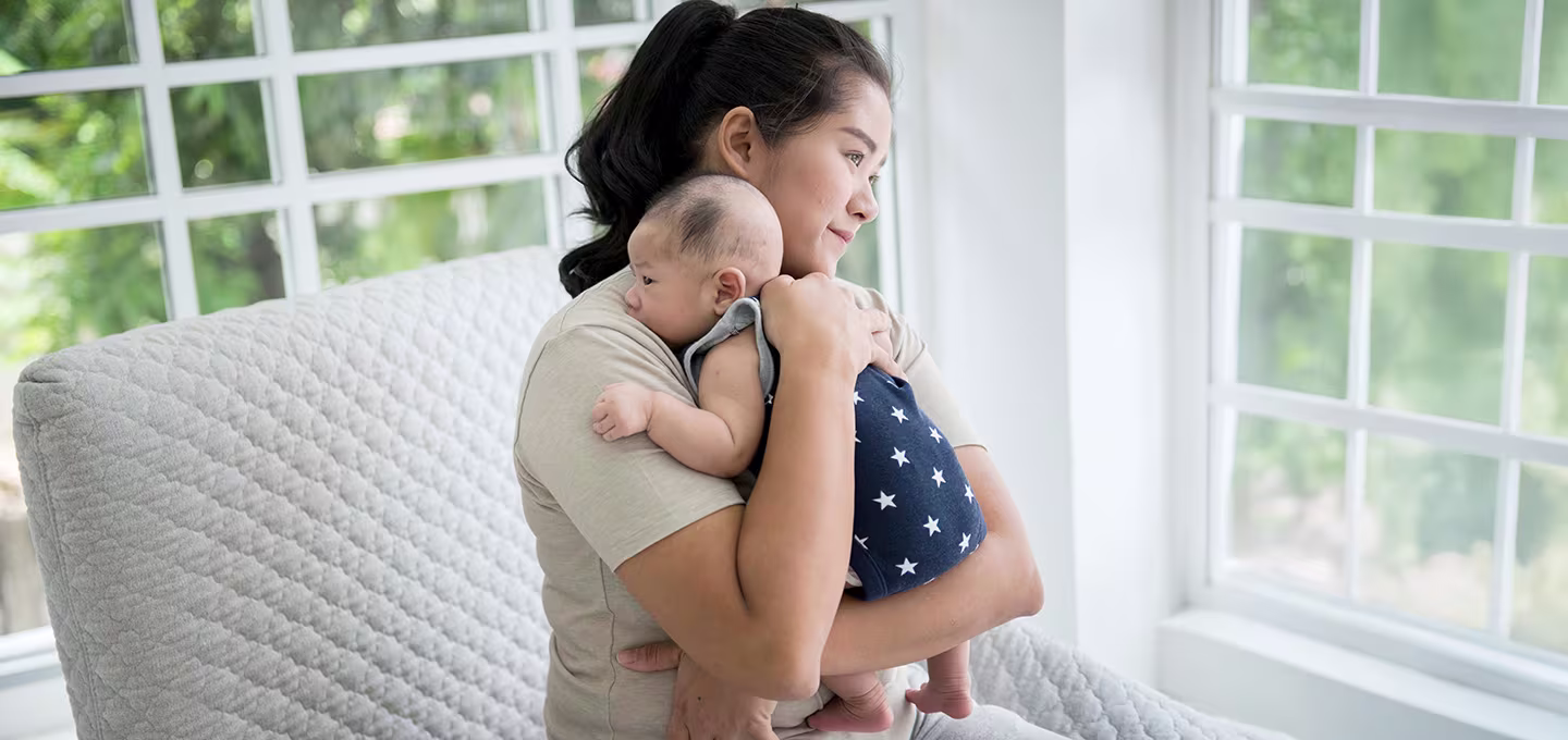 Mère tenant son bébé contre elle près d’une fenêtre lumineuse, le nourrisson reposant sur son épaule.