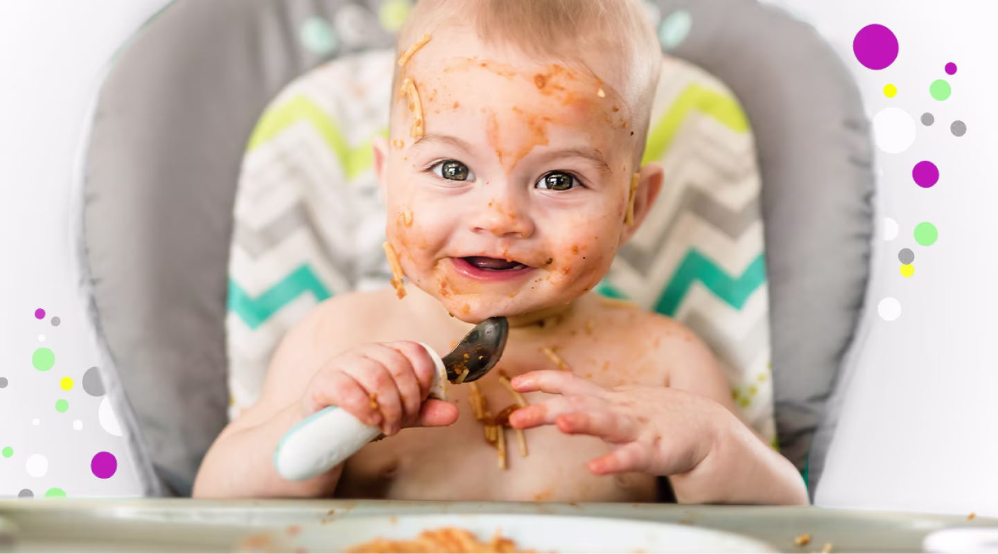 Smiling baby in a high chair, face covered in spaghetti sauce, holding a spoon