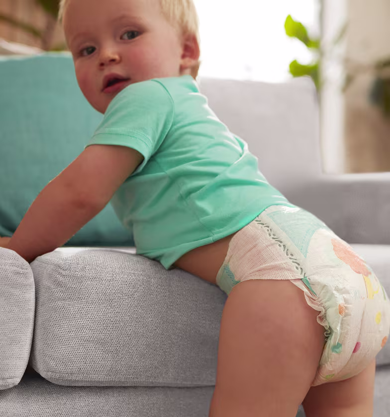 Bébé debout souriant, portant une couche Pampers, dans un environnement lumineux et doux évoquant le confort et la protection.
