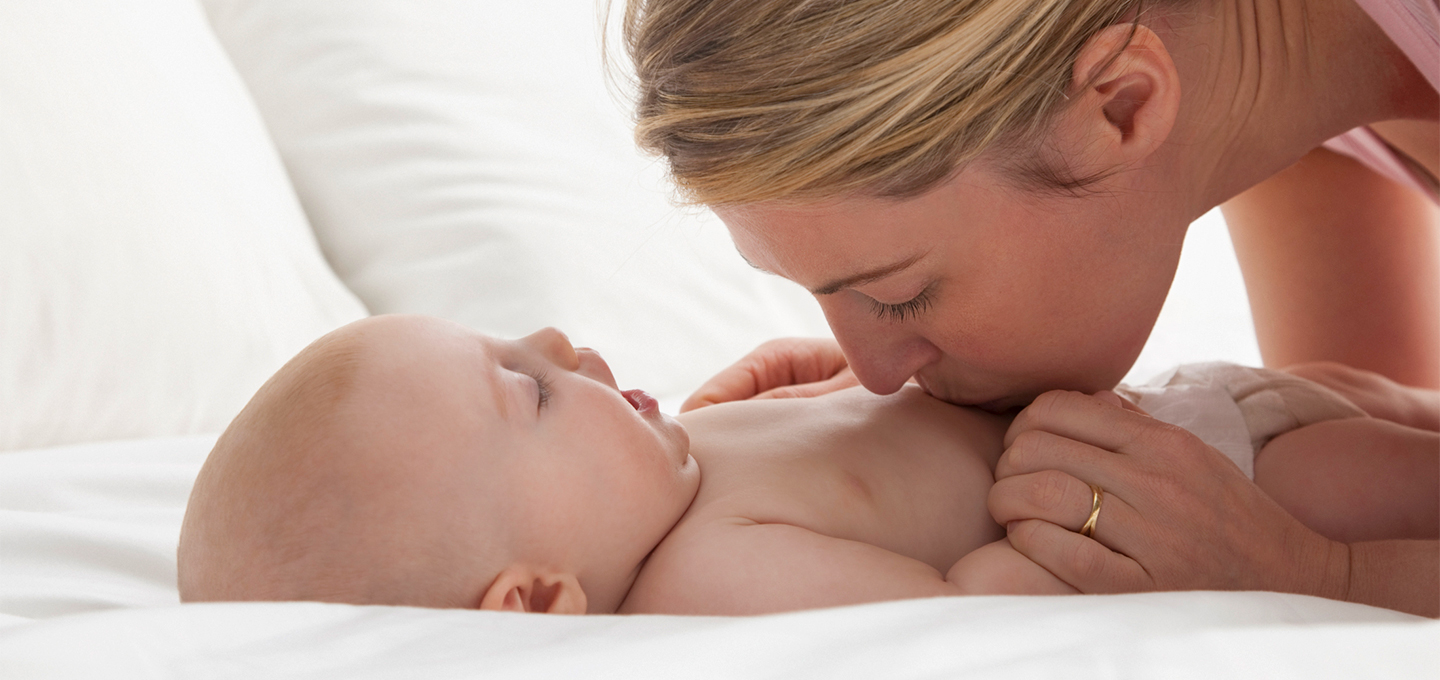 Mother lovingly kissing baby's tummy while the baby lies on a bed.