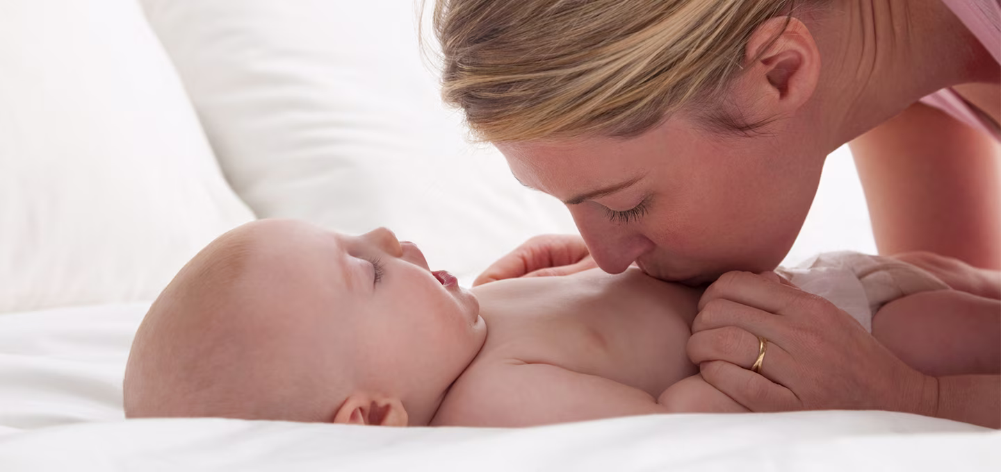 Mother lovingly kissing baby's tummy while the baby lies on a bed.