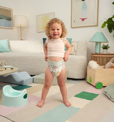 A smiling toddler standing in a colorful bright living room pulling up her Easy Ups training pant. A couch, artwork, lamp and potty in the background