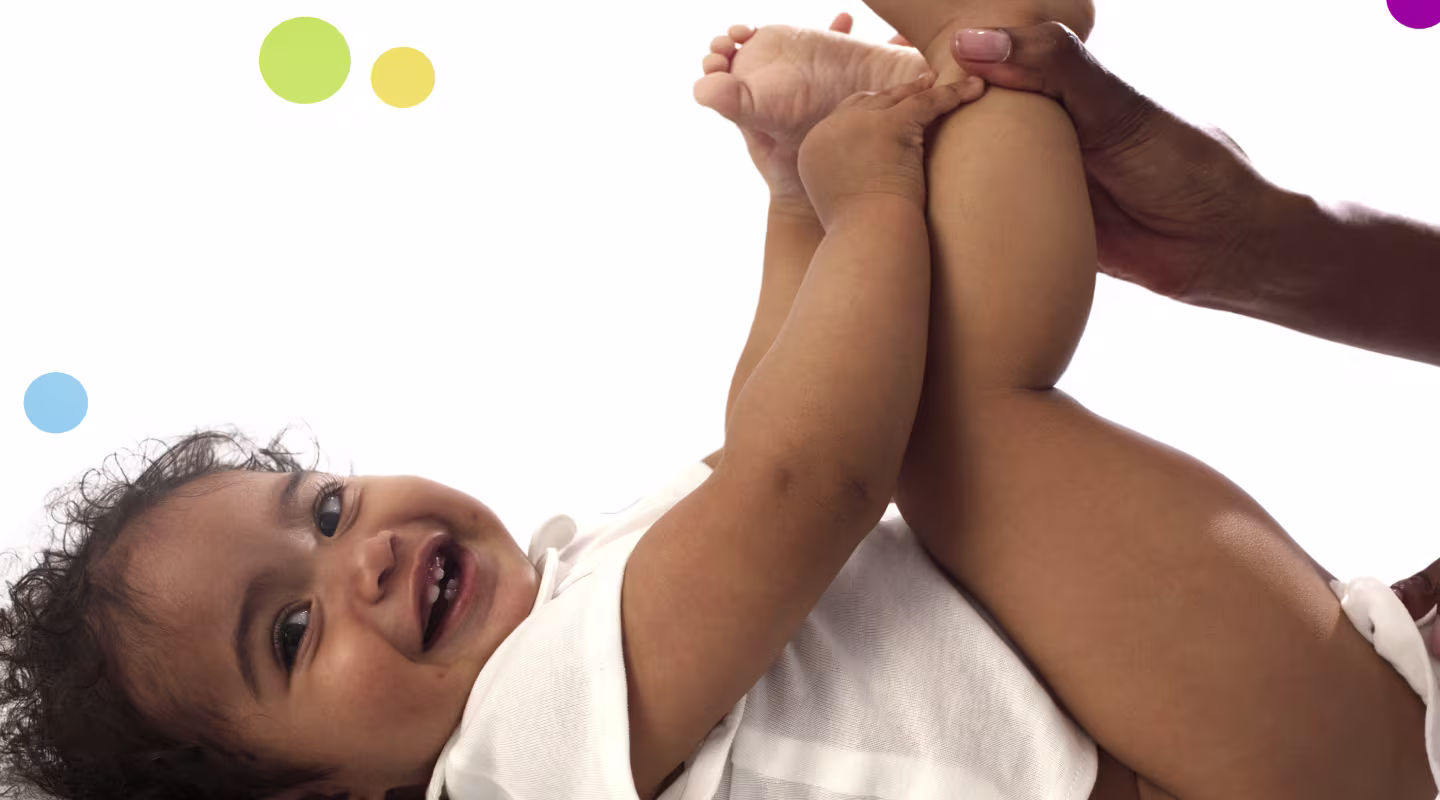 A baby lies on their back on a white surface, holding their feet up while a parent is using a wipe to clean the baby’s bottom.