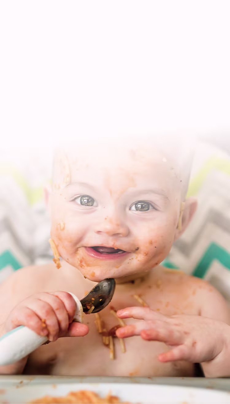Lächelndes Baby im Hochstuhl, Gesicht mit Spaghetti-Sauce bedeckt, hält einen Löffel