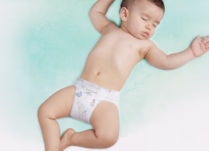 A baby sleeps soundly on their back, wearing a white nappy pant with blue bunny and carrot designs.