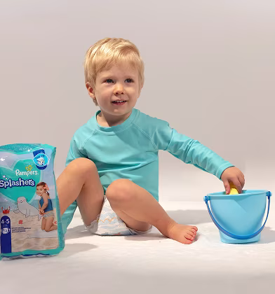 A cheerful baby wearing colorful Pampers Splashers, sitting with a bag of Splashers diapers and a bucket and spade
