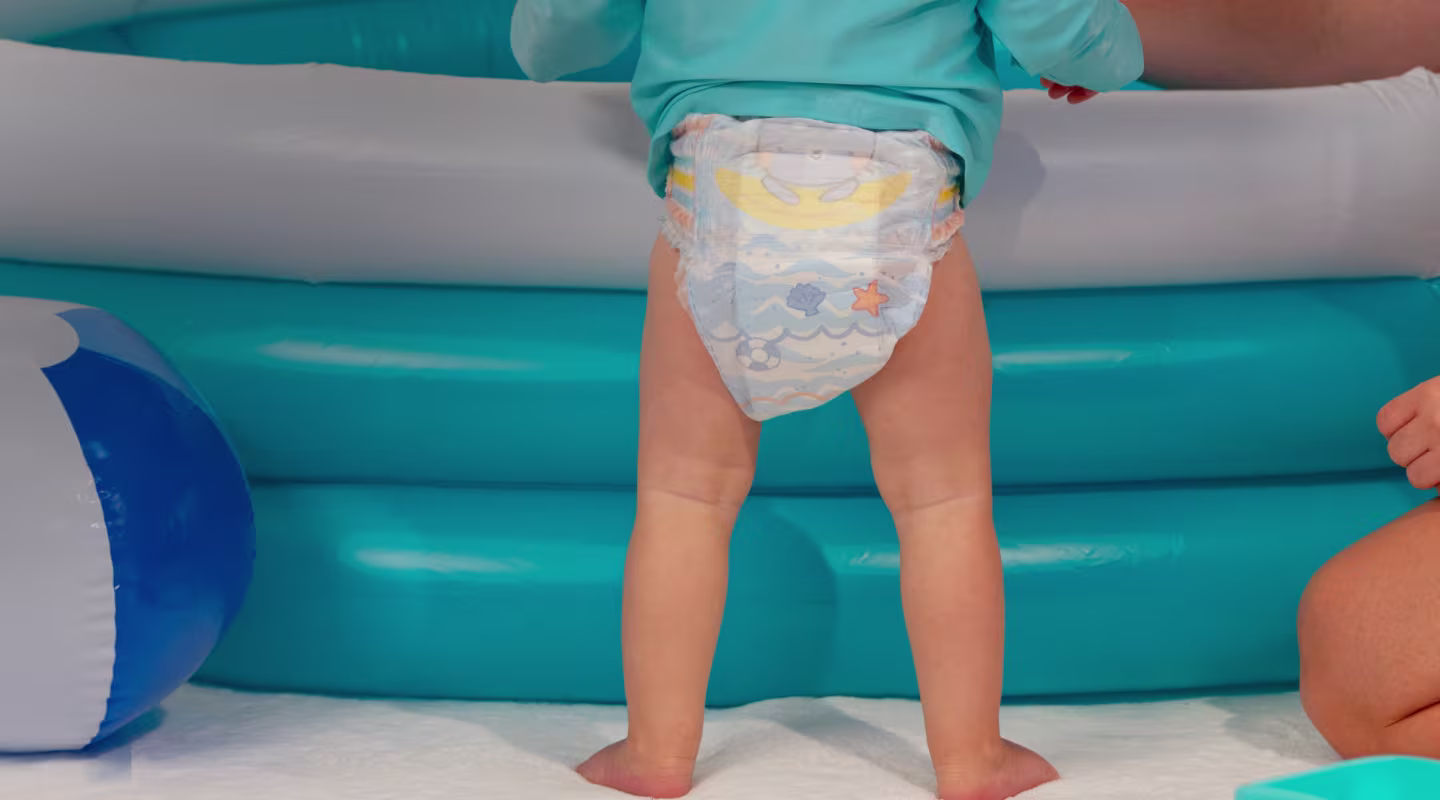 Un bebé alegre se encuentra de pie con confianza en la playa, luciendo sus coloridos Pampers Splashers, listo para un día de diversión bajo el sol.