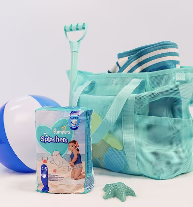 A beach bag sits packed with a towel, colorful toys, and sand shovel next to a beach ball and pack of Pampers Splashers