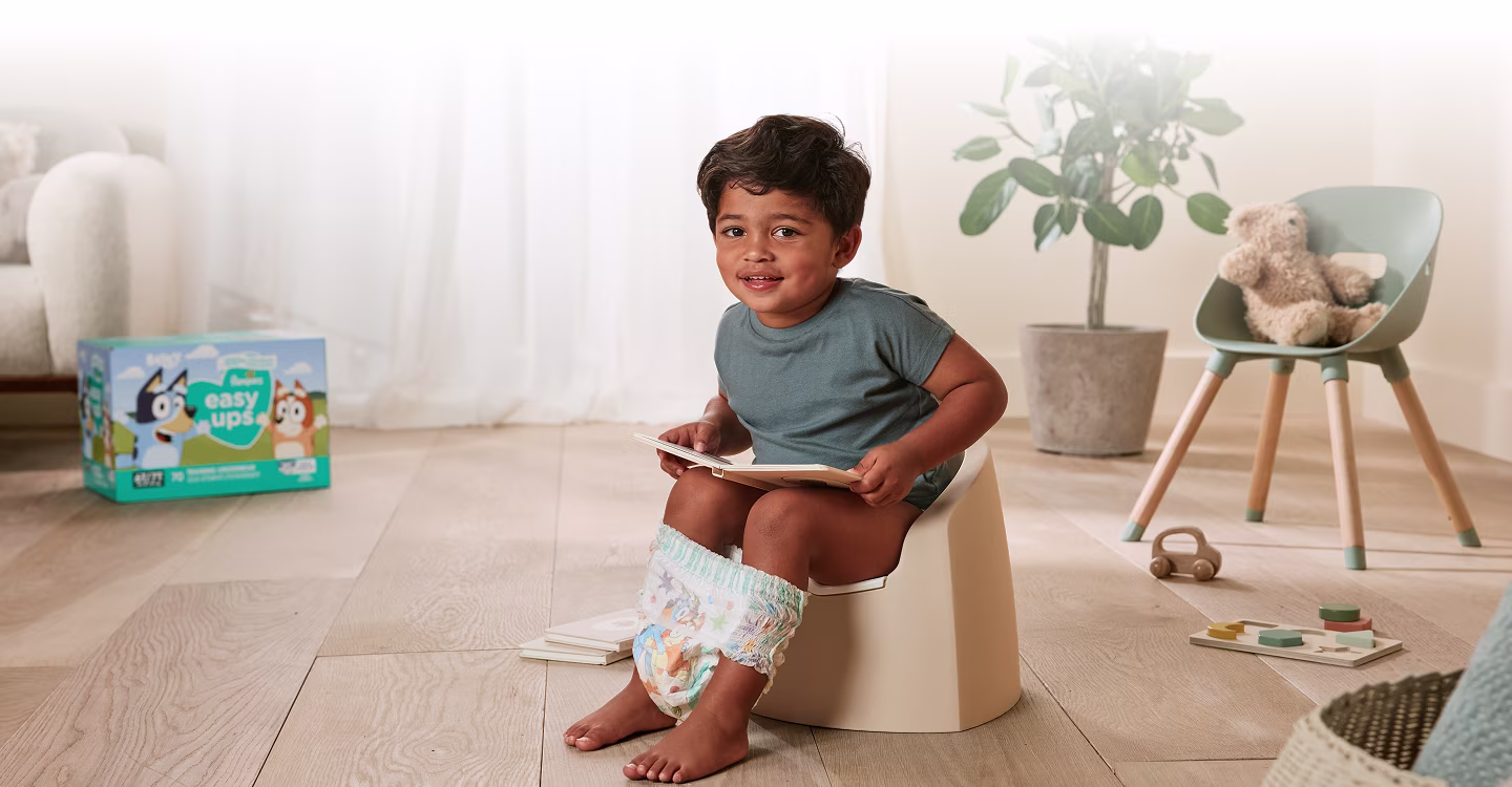 A toddler sits on a small, colorful potty chair in a brightly lit bathroom.