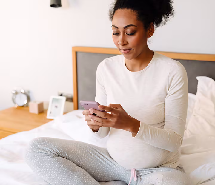Pregnant woman in comfy clothes sitting on bed, using smartphone.