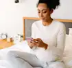 Pregnant woman in comfy clothes sitting on bed, using smartphone.