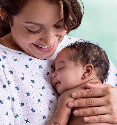 En smilende mamma i sykehusklær holder en sovende nyfødt baby tett inntil brystet sitt.