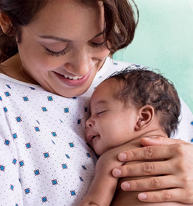 En smilende mamma i sykehusklær holder en sovende nyfødt baby tett inntil brystet sitt.