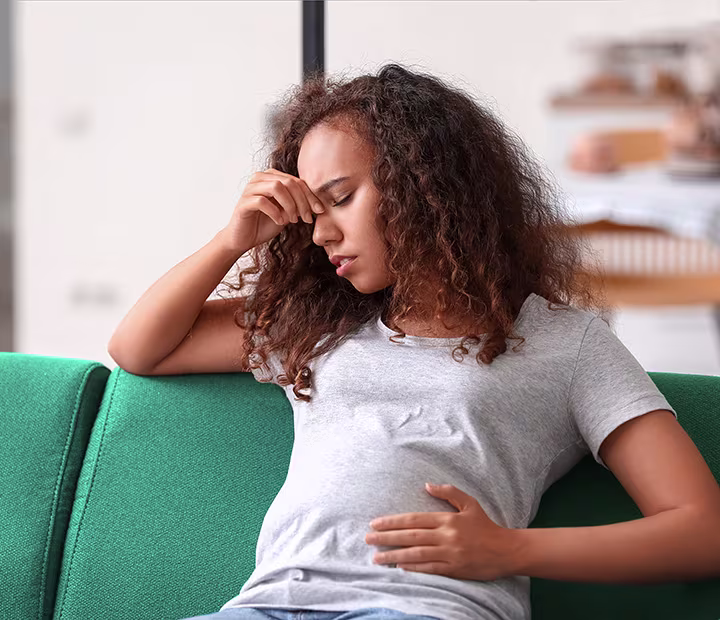 Pregnant woman sitting on a couch, looking concerned while touching her forehead and stomach. Pregnant woman sitting on a couch, looking concerned while touching her forehead and stomach.