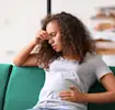 Pregnant woman sitting on a couch, looking concerned while touching her forehead and stomach.