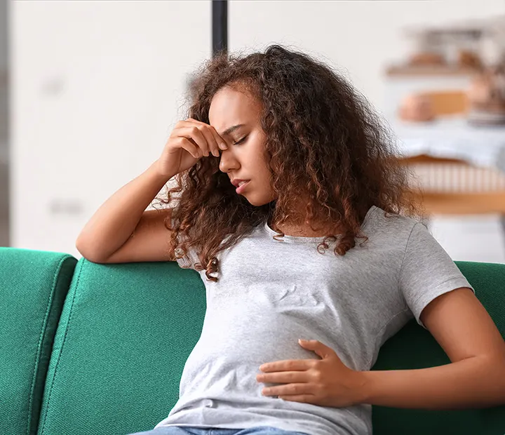 Pregnant woman sitting on a couch, looking concerned while touching her forehead and stomach.