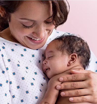 En leende mamma i sjukhuskläder håller en sovande nyfödd baby nära sitt bröst.