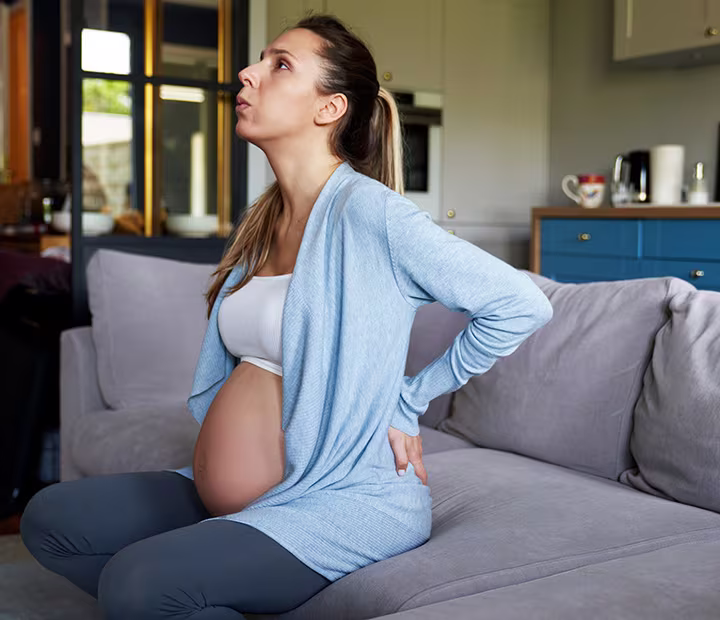 Pregnant woman in light blue cardigan sitting on sofa, holding back in discomfort. Pregnant woman in light blue cardigan sitting on sofa, holding back in discomfort.