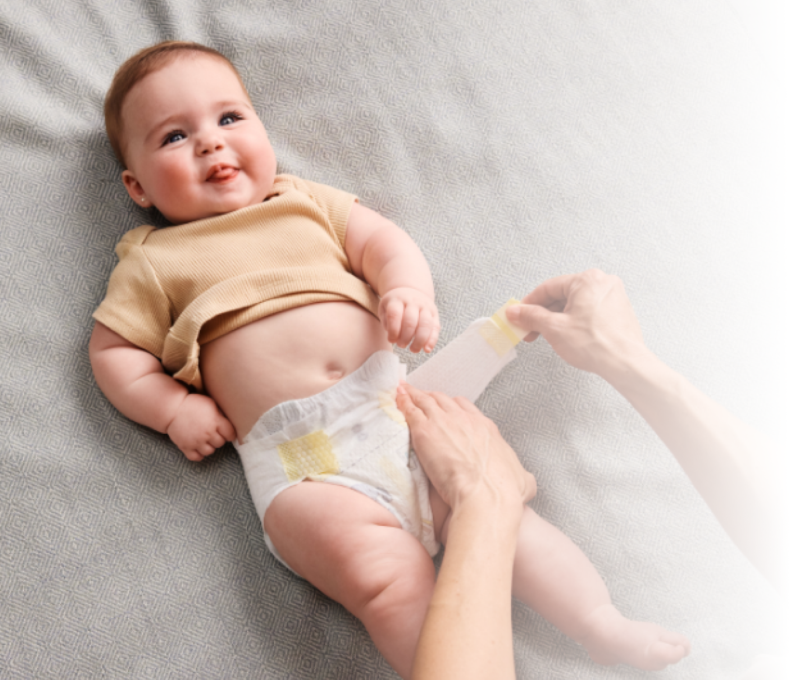 Baby smiling on bed get a diaper change, wearing Pampers Swaddlers. Mom’s hand showing the stretchy sides