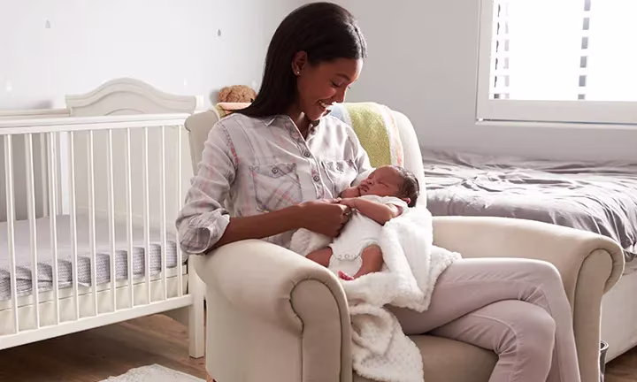 Eine Mutter sitzt vor einem Kinderbett auf einem Sessel und hält ihr Neugeborenes im Arm.
